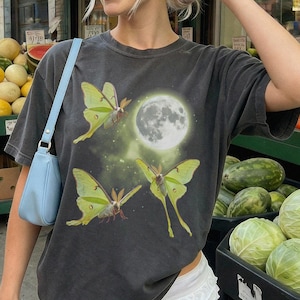 May include: A charcoal gray short-sleeve t-shirt featuring a graphic print of three lime green luna moths and a full moon against a dark background. The shirt is paired with white ruffled shorts and a light blue shoulder bag with a silver strap.
