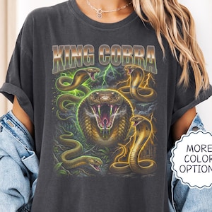 Op de afbeelding: Een antracietgrijs t-shirt met een grafische print van meerdere cobra's en de tekst KING COBRA. Het centrale ontwerp toont een grote cobra-kop met een open bek, omringd door kleinere slangen, groene bliksem en gele energie-effecten.