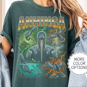 Peut inclure: Un t-shirt bleu épicéa avec une impression graphique représentant un oiseau Anhinga dans diverses poses, entouré d'éclairs verts et orange. Le texte ANHINGA est imprimé dans une grande police arquée en haut du motif graphique.