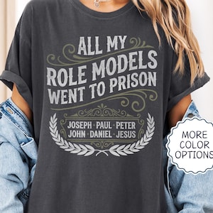 May include: A charcoal gray t-shirt featuring the text ALL MY ROLE MODELS WENT TO PRISON in a distressed white font, with a list of names including JOSEPH, PAUL, PETER, JOHN, DANIEL, and JESUS, framed by decorative flourishes and a laurel wreath.