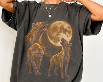 May include: A charcoal gray short-sleeve t-shirt featuring a graphic print of three hyenas in various poses beneath a large, glowing full moon. The design is rendered in shades of brown and gold, set against the dark fabric of the crew neck shirt.