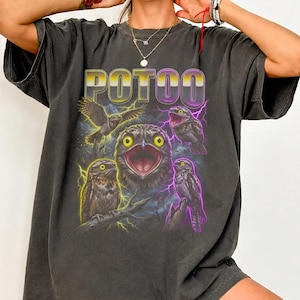 May include: A charcoal grey oversized t-shirt featuring the text POTOO in a yellow and purple gradient font. The graphic includes four illustrations of potoo birds with bright yellow eyes, surrounded by purple and yellow lightning bolts.