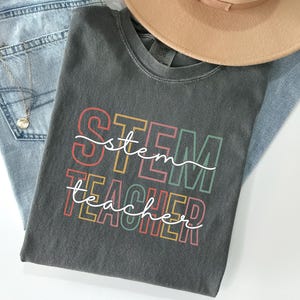 Peut inclure: Un t-shirt gris anthracite avec les mots "STEM TEACHER" dans un design coloré superposé. Le t-shirt est associé à un jean en denim délavé, un chapeau beige et un collier doré. Une brindille de fleurs séchées ajoute une touche décorative.