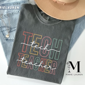 Pode incluir: Uma t-shirt cinza escuro com as palavras "TECH TEACHER" em letras coloridas e uma palavra "tech" em itálico. A t-shirt é combinada com jeans azul claro e um chapéu bege. A marca "MARIELAUREN" é visível nos jeans.