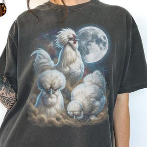 May include: Dark grey t-shirt featuring a whimsical design of three white chickens against a backdrop of a full moon and a starry sky. The chickens have fluffy feathers and are depicted in a surreal, artistic style. The shirt is made of a soft, comfortable material.