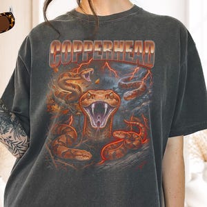 以下が含まれることがあります： メタリックなフォントでCOPPERHEADの文字がプリントされたチャコールグレーのTシャツ。デザインには、オレンジ色の稲妻と秋の葉に囲まれた、様々なポーズをとる複数のカッパーヘッドスネークが描かれています。