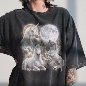 May include: Dark gray t-shirt featuring a fantasy design with three white unicorns and a full moon. One unicorn is rearing up, another is lying down, and a third is in a halo. The design is set against a cloudy background.