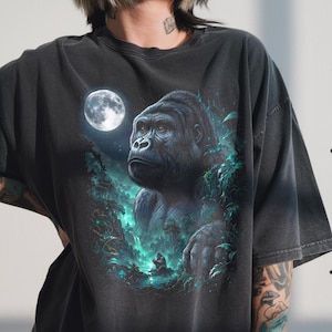 Puede incluir: Camiseta gris oscuro con una ilustración detallada de un gorila en un entorno selvático bajo la luna llena. La obra de arte incluye un exuberante follaje verde y una cascada, creando un diseño vibrante con temática natural.