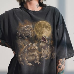 May include: A charcoal gray short-sleeve t-shirt featuring a graphic print of three textured brown toads positioned beneath a large, glowing full moon. The design includes hazy, golden-brown atmospheric clouds surrounding the toads and the moon.