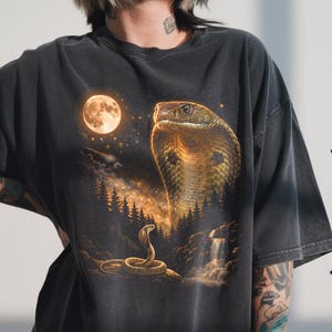 May include: Dark grey t-shirt with a detailed illustration of a cobra in a moonlit forest. The design includes a full moon, a waterfall, and a second cobra. The cobra's head is prominently displayed, with a detailed scale pattern.