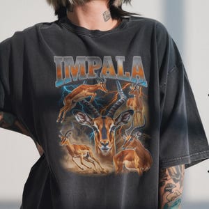 以下が含まれることがあります： インパラのグラフィックとオレンジと青の「IMPALA」の文字が入った濃いグレーのTシャツ。デザインには、背景に稲妻のある複数のインパラが含まれています。