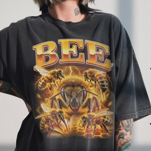 以下が含まれることがあります： 大きな蜂のグラフィックと「BEE」の文字が金色で描かれた濃い灰色のTシャツ。グラフィックには複数の蜂とハニカムデザインが描かれています。