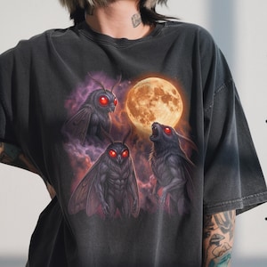 Può includere: T-shirt grigio scuro con una grafica di tre creature simili a falene con occhi rossi e un lupo che ulula alla luna piena. Il design è ambientato su uno sfondo di nuvole viola e una luna arancione brillante.