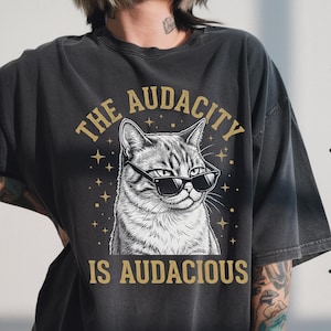May include: Dark gray t-shirt featuring a detailed illustration of a cat wearing sunglasses. The text "THE AUDACITY IS AUDACIOUS" is printed in gold above and below the cat image. The shirt has a vintage, worn look.