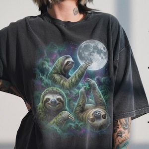 May include: Dark grey t-shirt with a fantasy design of three sloths and a full moon. The sloths are in green and brown, set against a swirling purple and green nebula. A unique and eye-catching design.