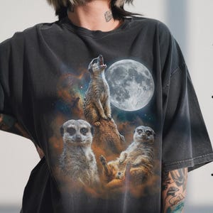 May include: Dark gray t-shirt featuring a graphic of meerkats under a full moon. The design includes a meerkat howling at the moon, with two others below. The background has a nebula effect.