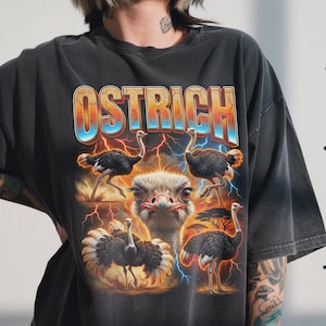 May include: Dark gray t-shirt featuring a graphic of ostriches with the word "OSTRICH" in a colorful, retro font. The design includes multiple ostrich illustrations, lightning bolts, and a savanna landscape.