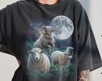 Puede incluir: Camiseta gris oscuro con un diseño humorístico de tres ovejas en un entorno cósmico. Una oveja salta, otra lleva un sombrero de cubo y una tercera mira hacia adelante. La luna en el fondo tiene la palabra "BAAAA".