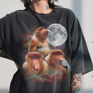 May include: Dark gray t-shirt featuring a graphic of three proboscis monkeys with a full moon and a nebula background. The monkeys have reddish-brown fur and large noses. The shirt has a vintage, worn look.