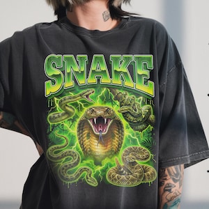 Könnte beinhalten: Dunkelgraues T-Shirt mit einem lebendigen Grafikdesign. Das Wort "SNAKE" ist in Grün und Gelb gehalten. Das Design zeigt mehrere Schlangen, Blitze und eine Kobra mit offenem Maul, was einen kühnen, kantigen Stil zeigt.