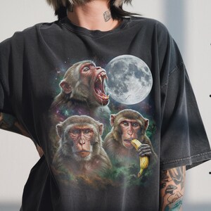 May include: Dark gray t-shirt featuring a vibrant graphic of three monkeys against a cosmic background with a full moon. One monkey is eating a banana, another is screaming, and the third is looking forward.