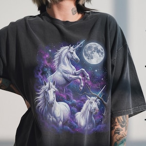 May include: Dark gray t-shirt featuring a fantasy design with three white unicorns against a backdrop of a full moon, stars, and swirling purple and blue nebula. The unicorns have detailed manes and horns, creating a mystical scene.