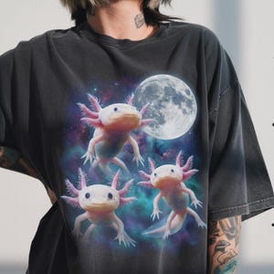May include: Dark grey t-shirt with a graphic of three axolotls in space, a full moon behind them. The axolotls are pink and white with feathery gills. The background is a mix of purple, blue, and green.