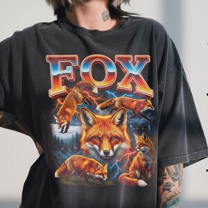 A vintage washed black t-shirt featuring a Fox Bootleg 90s Vintage Graphic. The design displays bright red foxes hunting and walking in a dark misty forest with glowing eyes in the background, lightning effects, and retro blue-orange gradient typography.
