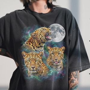 Puede incluir: Camiseta gris oscuro con un gráfico de tres leopardos y una luna llena. Los leopardos tienen manchas amarillas y negras y ojos verdes. El fondo presenta un diseño colorido similar a una galaxia.