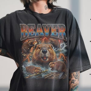 May include: Dark gray t-shirt featuring a graphic of beavers in a water scene with lightning bolts. The word "BEAVER" is printed across the top in a colorful, outlined font. The shirt has a vintage, distressed look.