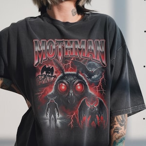 Può includere: T-shirt grigio scuro con una grafica di Mothman con occhi rossi, ali e la scritta "MOTHMAN" in rosso e bianco. Il design include un ponte, fulmini e una luna piena. La maglietta è in stile vintage.
