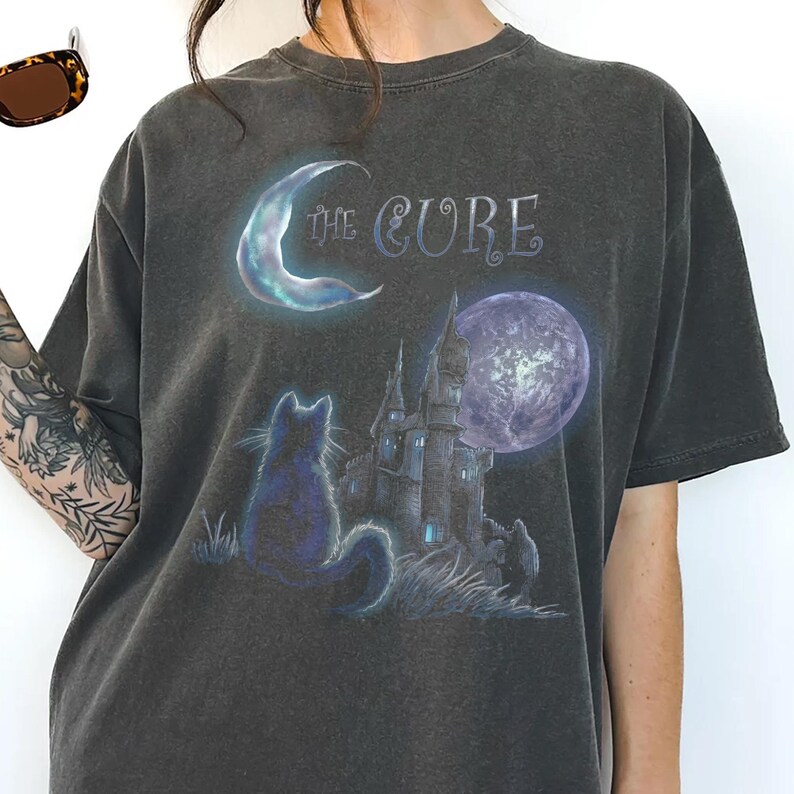 Dark grey tshirt featuring gothic text The Cure with a silhouette black cat sitting in front of a dark castle and full moon.