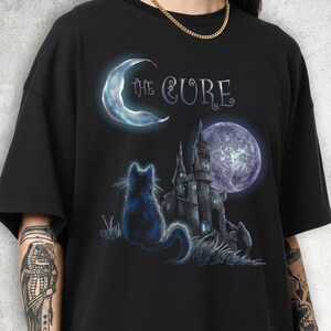 Dark grey tshirt featuring gothic text The Cure with a silhouette black cat sitting in front of a dark castle and full moon.