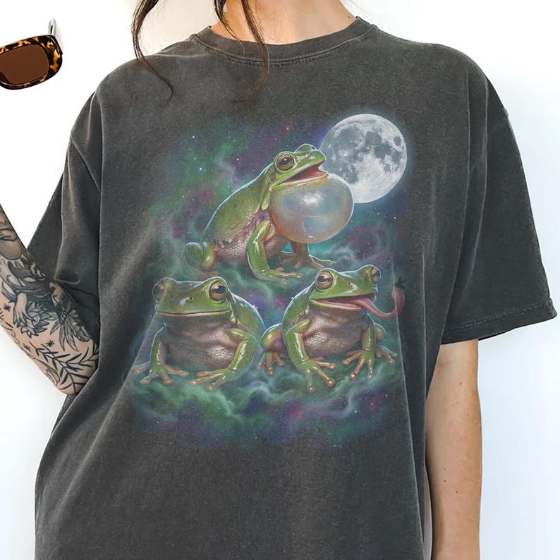 Vintage style three green tree frogs moon graphic printed on dark washed t-shirt, croaking frog with inflated throat and frog catching fly galaxy design.