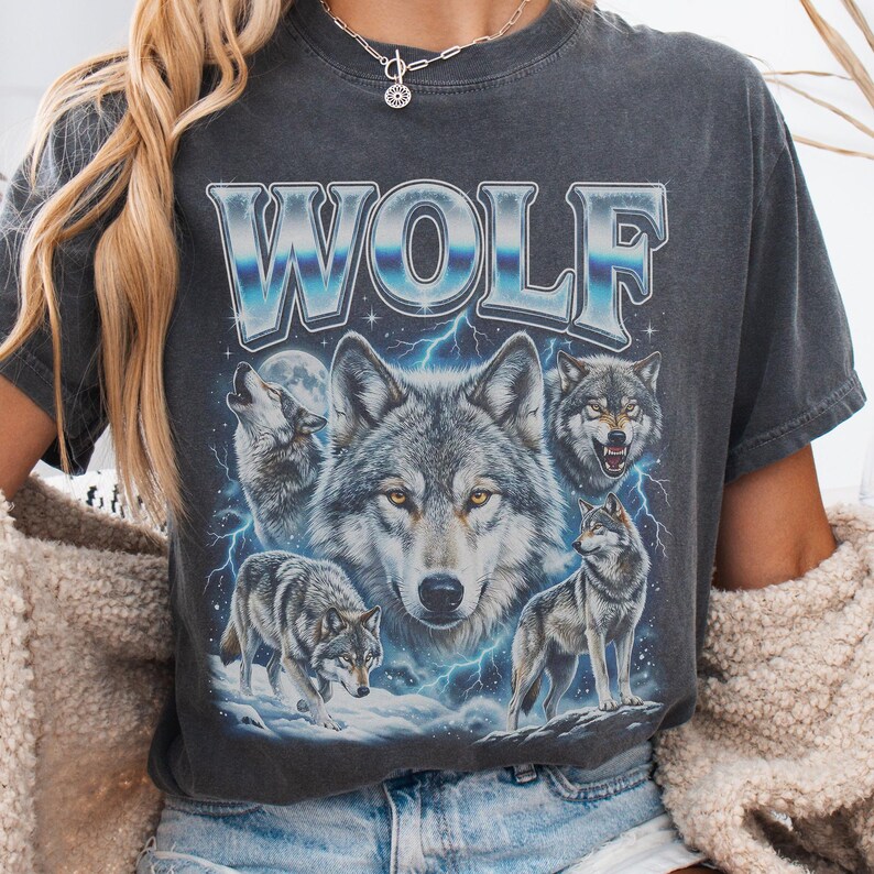 Model wearing a vintage wash black t-shirt featuring a collage of wolves howling at the moon with icy blue lightning, large metallic text reading WOLF in 90s bootleg style.