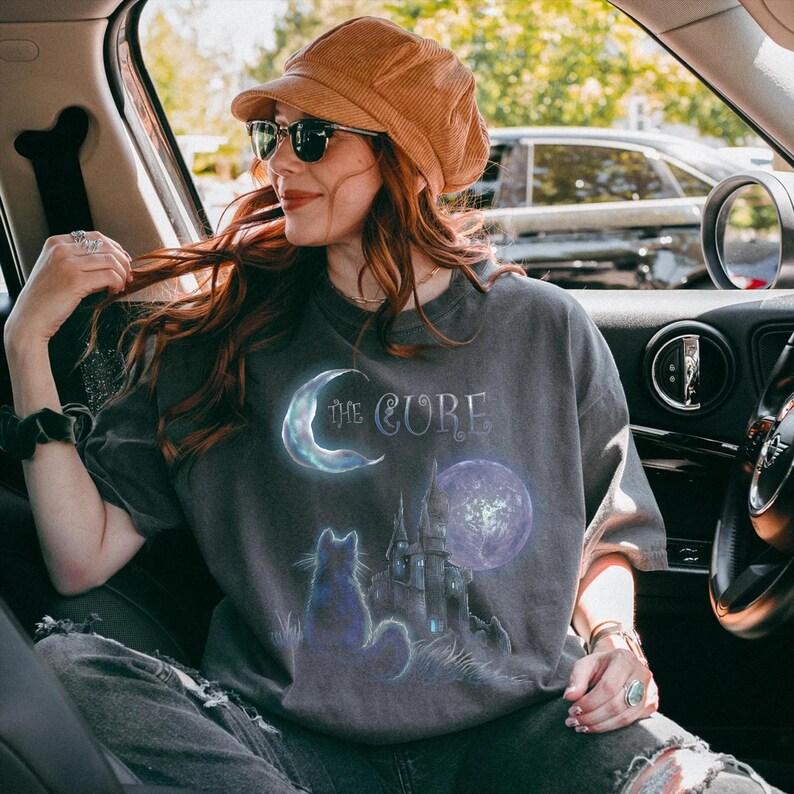 Dark grey tshirt featuring gothic text The Cure with a silhouette black cat sitting in front of a dark castle and full moon.