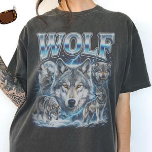 Model wearing a vintage wash black t-shirt featuring a collage of wolves howling at the moon with icy blue lightning, large metallic text reading WOLF in 90s bootleg style.