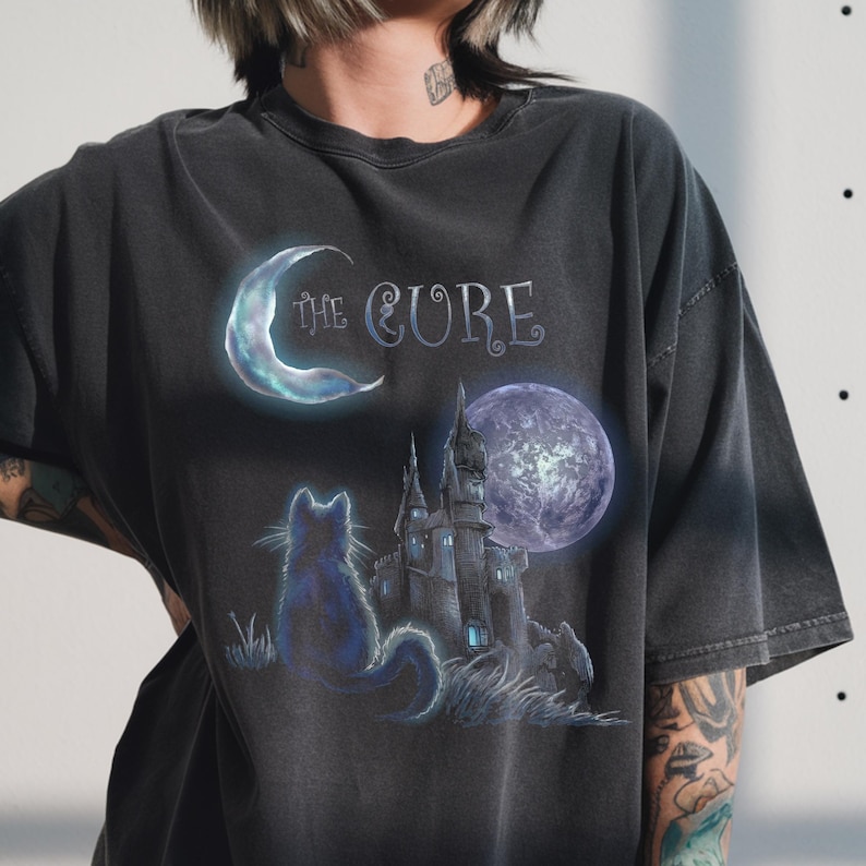Dark grey tshirt featuring gothic text The Cure with a silhouette black cat sitting in front of a dark castle and full moon.
