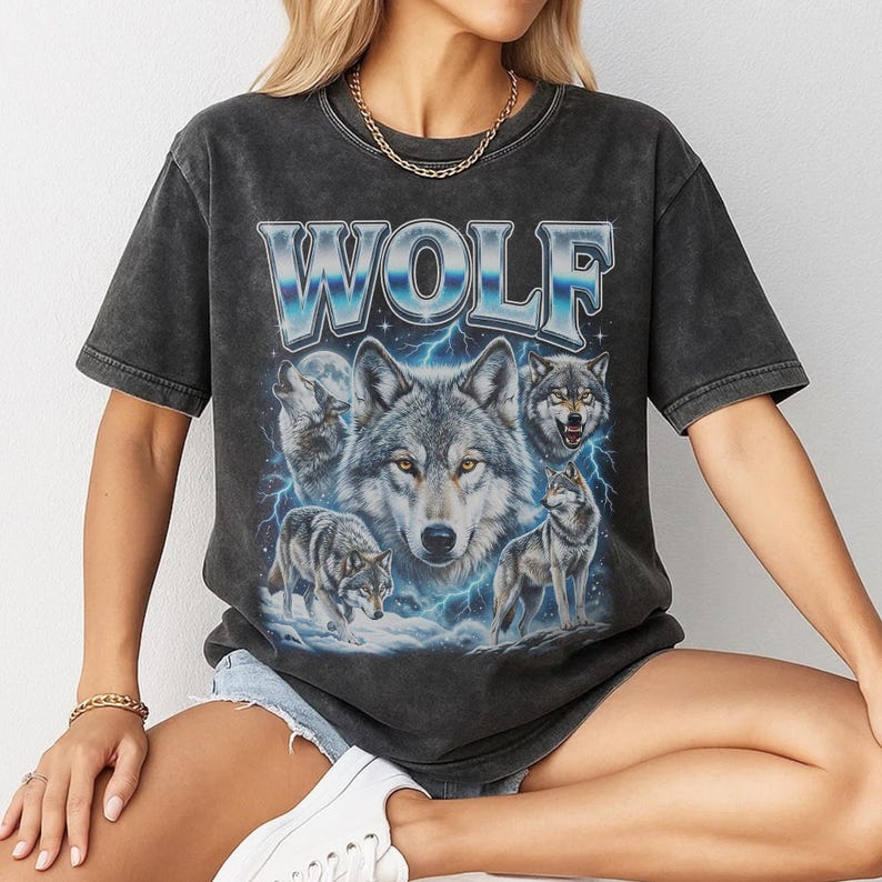 Model wearing a vintage wash black t-shirt featuring a collage of wolves howling at the moon with icy blue lightning, large metallic text reading WOLF in 90s bootleg style.