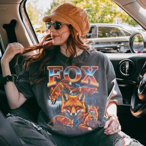 May include: Dark gray t-shirt with a graphic of several foxes and the word "FOX" in a retro font. The foxes are depicted in shades of orange and brown, with lightning bolts in the background. The shirt is worn by a person wearing sunglasses and a brown hat.