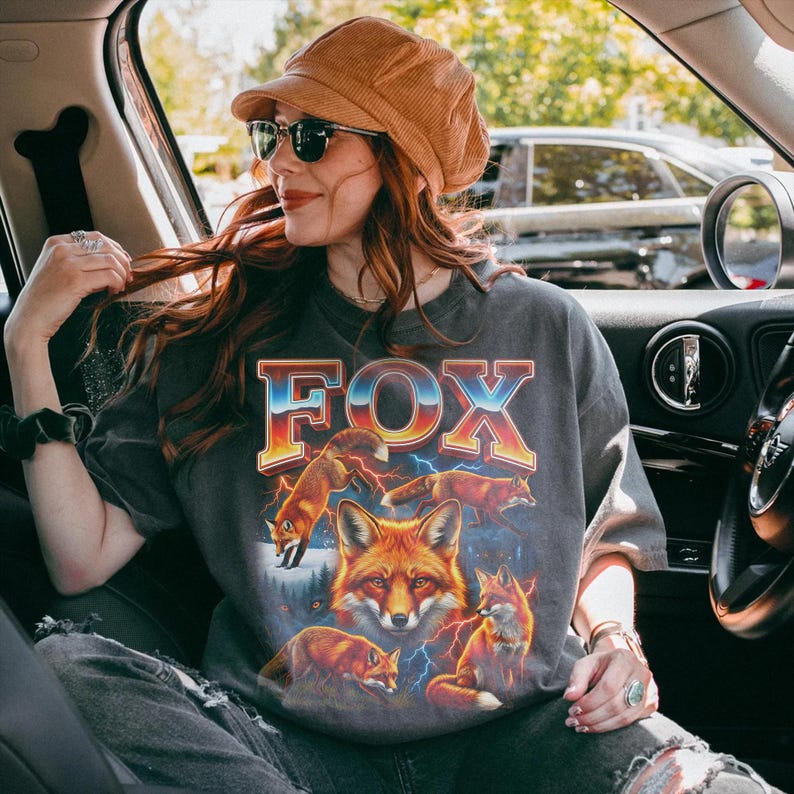 May include: Dark gray t-shirt with a graphic of several foxes and the word "FOX" in a retro font. The foxes are depicted in shades of orange and brown, with lightning bolts in the background. The shirt is worn by a person wearing sunglasses and a brown hat.