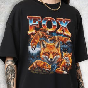 May include: Black t-shirt featuring a vibrant graphic of multiple foxes, with the word "FOX" in a retro font. The design includes orange and white foxes, lightning bolts, and a forest scene. The shirt is made of a soft material.