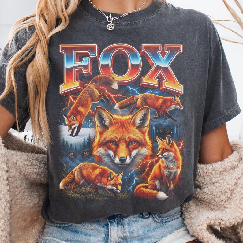May include: Dark gray t-shirt featuring a vibrant graphic of foxes and the word "FOX" in a colorful, retro font. The design includes multiple foxes, lightning bolts, and a forest scene.