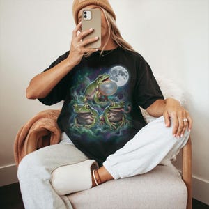 Vintage style three green tree frogs moon graphic printed on dark washed t-shirt, croaking frog with inflated throat and frog catching fly galaxy design.