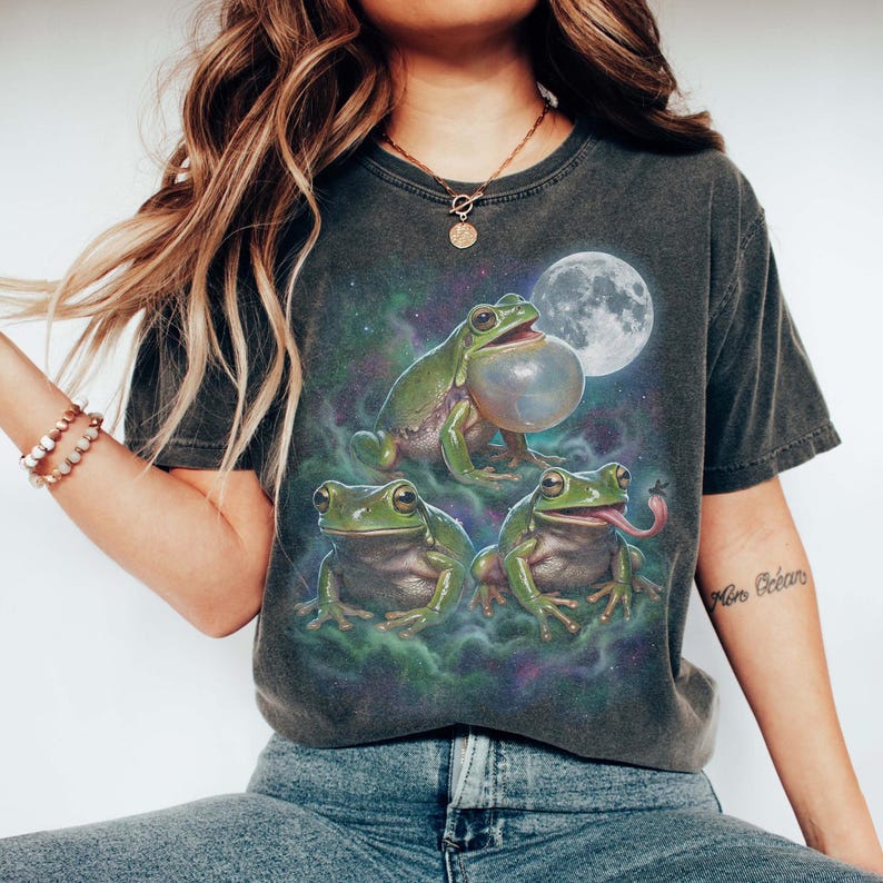 Vintage style three green tree frogs moon graphic printed on dark washed t-shirt, croaking frog with inflated throat and frog catching fly galaxy design.