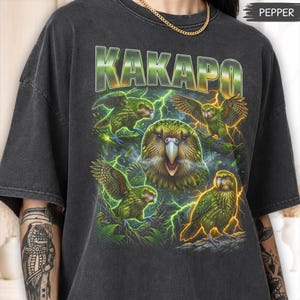 Puede incluir: Una camiseta gris carbón con un estampado gráfico de varios loros kakapo verdes rodeados de rayos amarillos. La palabra KAKAPO está impresa en letras grandes, negritas y de estilo metálico en la parte superior. La camiseta tiene cuello redondo.