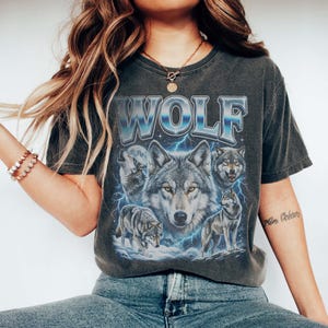 Model wearing a vintage wash black t-shirt featuring a collage of wolves howling at the moon with icy blue lightning, large metallic text reading WOLF in 90s bootleg style.