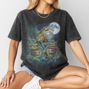 Vintage style three green tree frogs moon graphic printed on dark washed t-shirt, croaking frog with inflated throat and frog catching fly galaxy design.