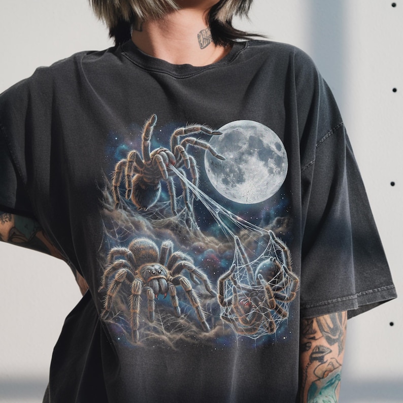 Three realistic tarantula spiders under a large luminous full moon in a starry night sky. One spider at the top spins a web toward the moon. Below, one spider faces forward with funny googly eyes, while the third is entangled in silk webbing. The design features a retro 90s galaxy aesthetic with cosmic nebula clouds.