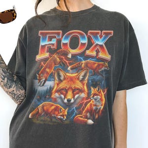 May include: Dark gray t-shirt featuring a vibrant graphic of foxes and the word "FOX" in a retro font. The design includes multiple foxes, lightning bolts, and a forest scene, creating a vintage, outdoorsy aesthetic.